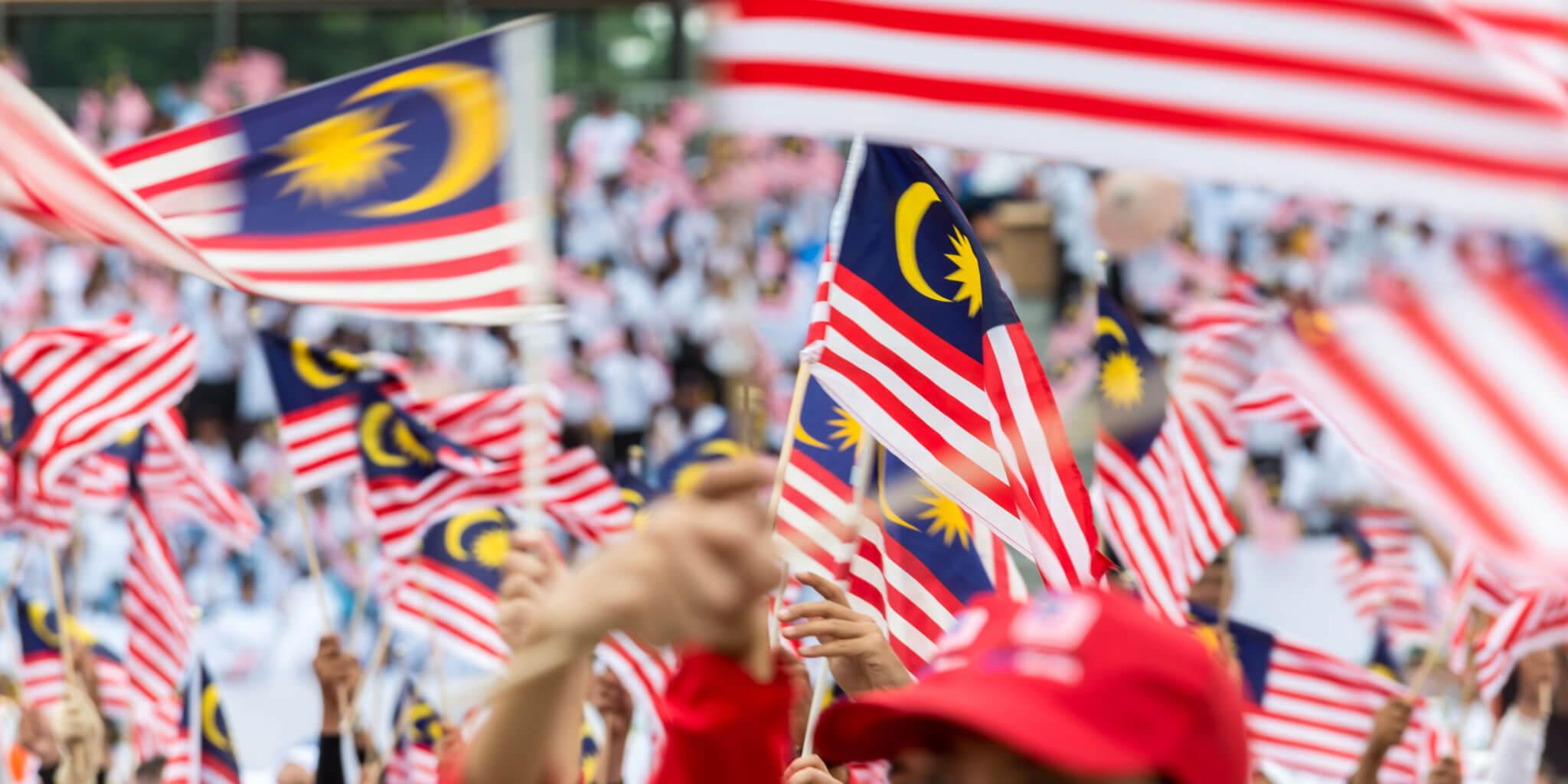 A crowd of people waving multiple Malaysian flags. The flags display red and white stripes with a blue field in the upper left corner containing a yellow crescent moon and star. The crowd is blurred in the background, focusing on the flags in the foreground.