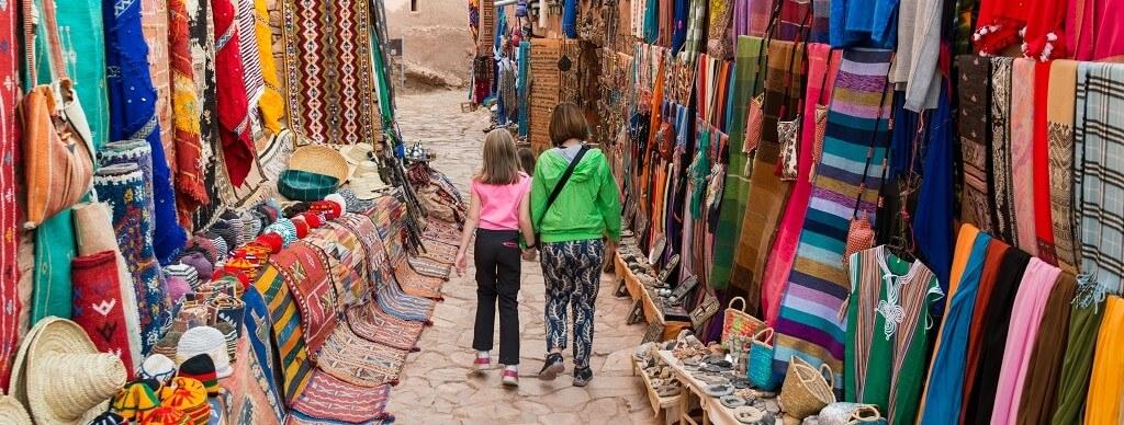 Two people, possibly a mother and daughter, walk hand in hand through a vibrant outdoor market in Morocco. The cobblestone path is lined with colorful textiles, rugs, and various other goods. The market stalls are densely packed with items on both sides, reflecting the rich tapestry of migration and remittances.