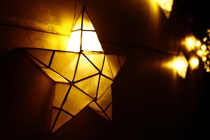 A glowing star-shaped lantern hangs on a wall, illuminated warmly in the evening. Behind it, more star-shaped lanterns glow softly, creating a cozy and festive ambiance. The dark background emphasizes the warm light emitted from the lanterns.