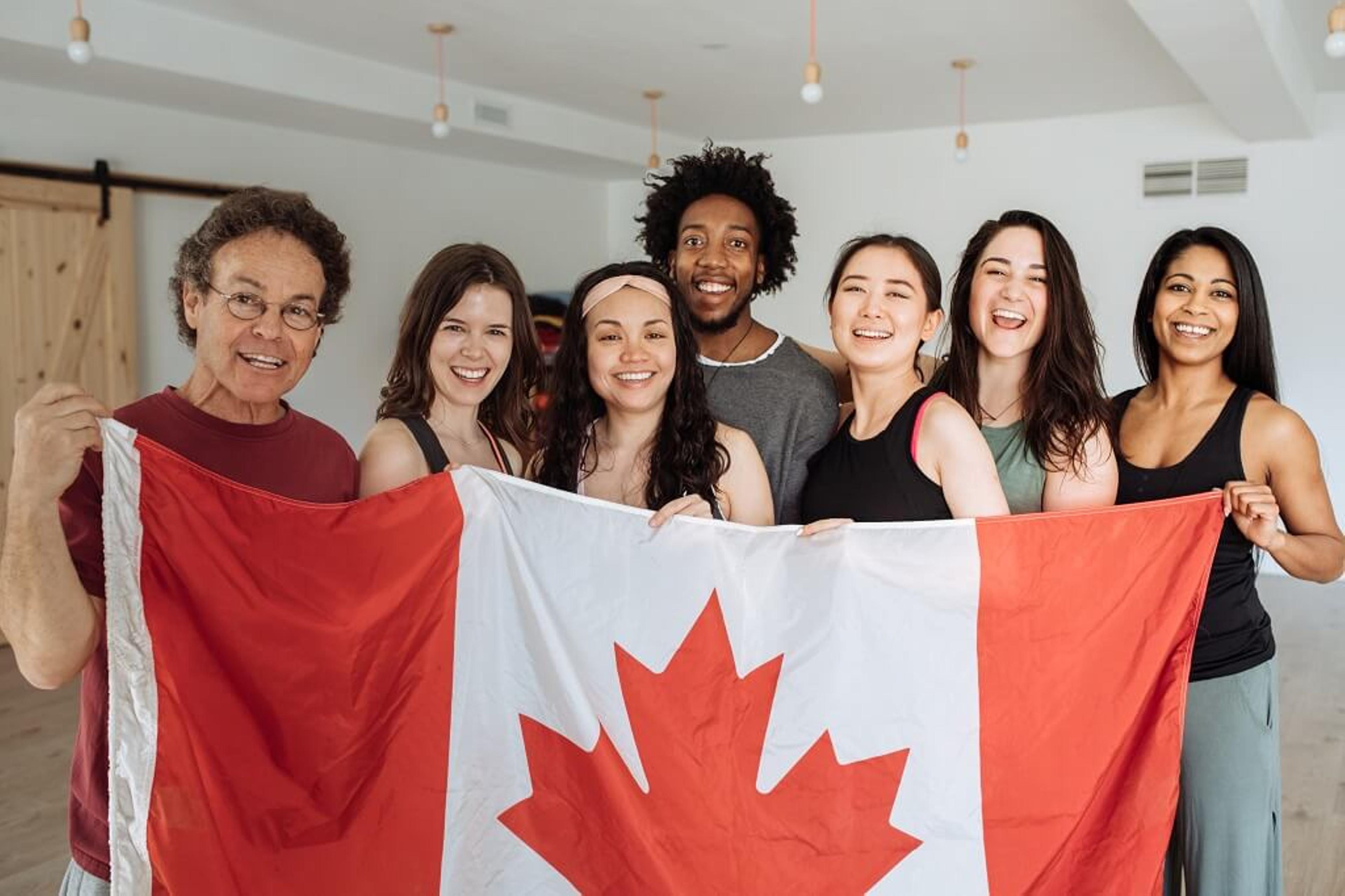 A diverse group of seven smiling people stands close together, holding a large Canadian flag. They are in a bright room with light-colored walls and wooden accents. The atmosphere appears joyful and celebratory, reflecting the positive impacts of immigration in Canada.