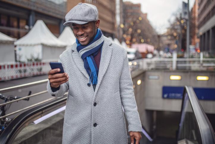 A person wearing a gray flat cap, gray coat, and blue scarf is smiling while looking at their phone. They are on an outdoor escalator in an urban area with buildings and tents in the background, possibly checking how to send money to Ghana from the United States. The weather appears chilly.