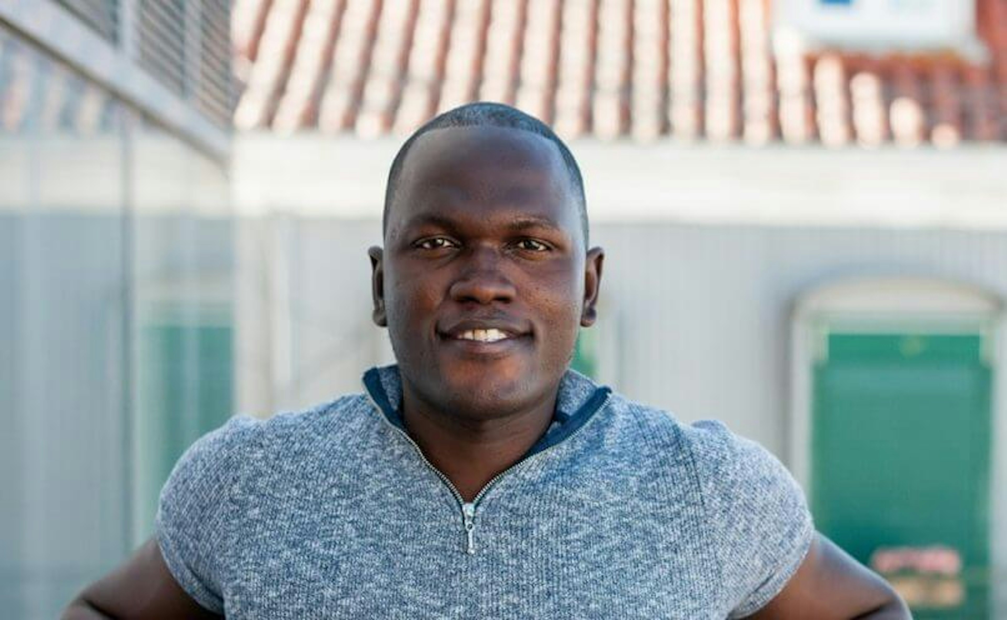 A person wearing a grey short-sleeve shirt stands outdoors with a building and a tiled roof in the background. They are looking directly at the camera, smiling slightly, with their hands on their hips, radiating cool confidence like they just stepped out of a Saico ad.