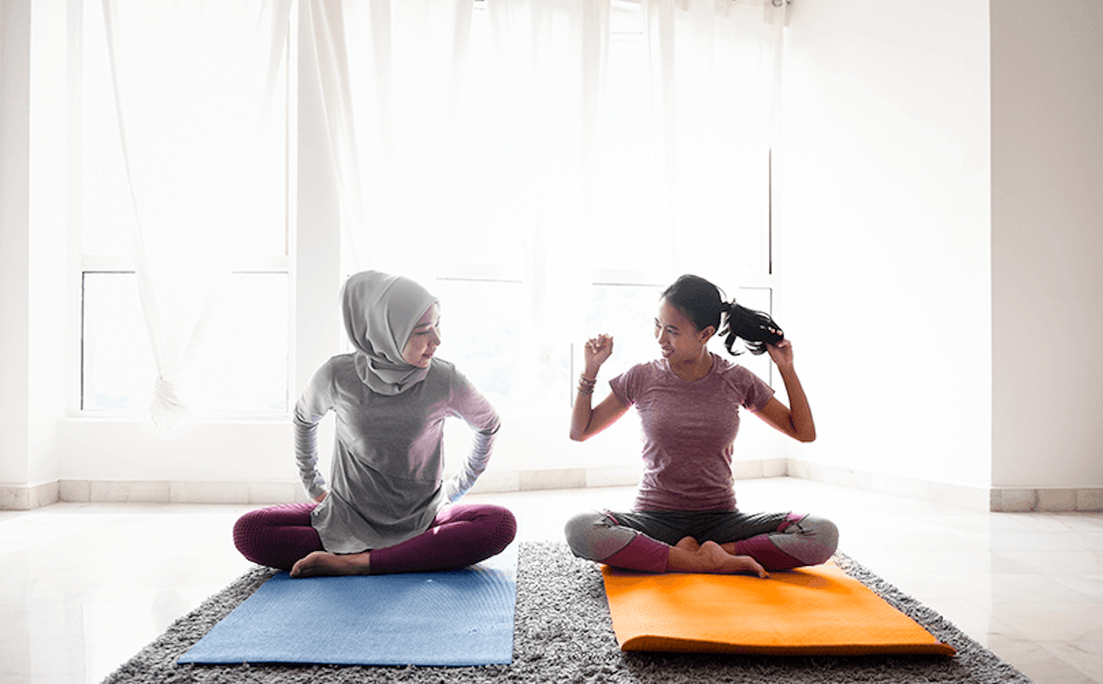 Dos mujeres se sientan sobre colchonetas de yoga en una habitación luminosa con cortinas transparentes. Una lleva un hiyab y la otra tiene el pelo recogido en una cola de caballo. Ambos parecen estar estirándose y sonriéndose el uno al otro. La mujer de la izquierda se sienta sobre una alfombra azul y la mujer de la derecha se sienta sobre una alfombra naranja.