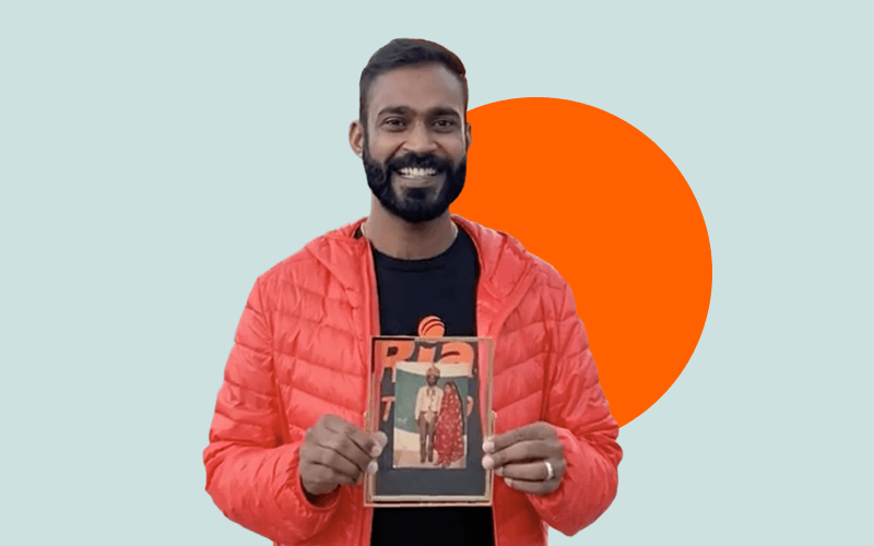 A man in a red quilted jacket and black shirt smiles while holding up a framed photograph of two people. The background is light blue with an orange circle behind him.