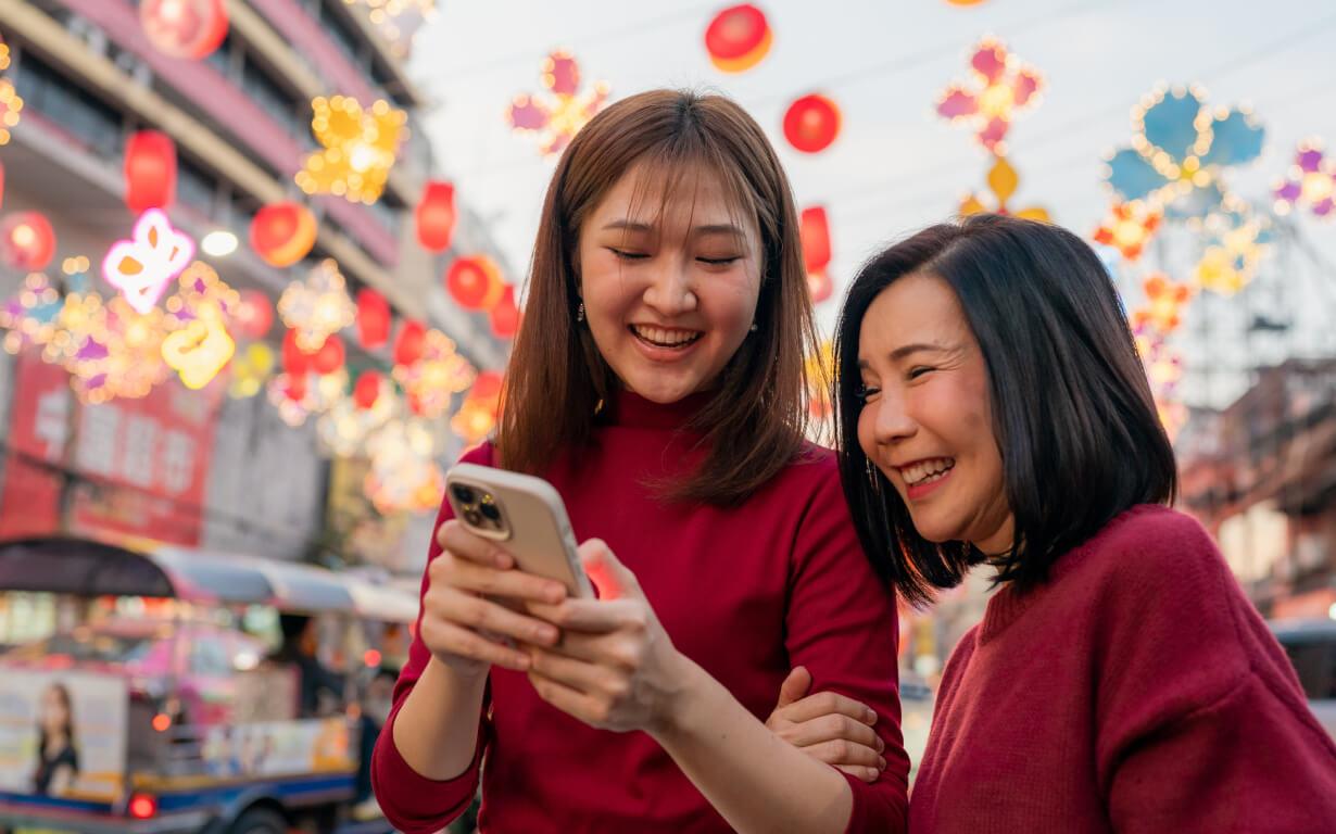 Dos mujeres con atuendos festivos sonríen mientras miran un teléfono inteligente, celebrando el Año Nuevo Lunar. Están de pie en una calle concurrida adornada con faroles y luces de colores. Se ven edificios y un vehículo en el fondo, lo que realza la atmósfera vibrante de la celebración.