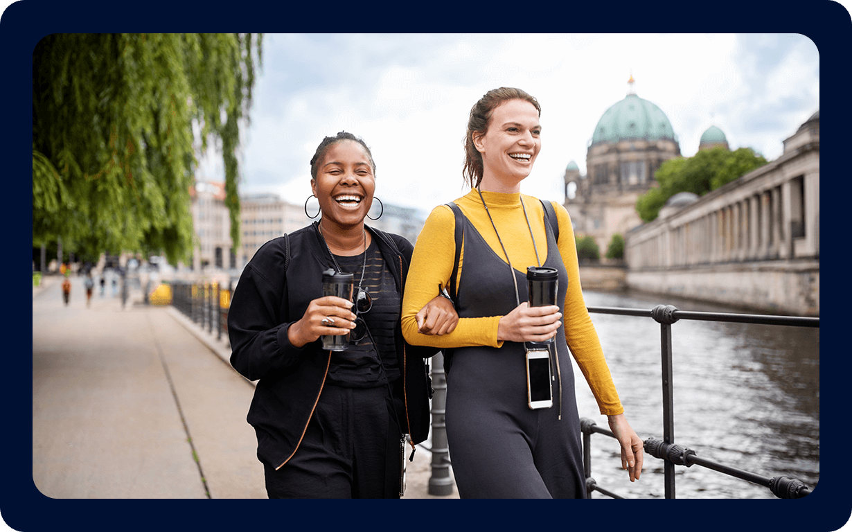 Two women walking arm-in-arm along a riverside path, laughing and holding takeaway coffee cups. One woman is wearing a black outfit and the other wearing a grey dress with a yellow top. In the background, a large domed building and bridge are visible.