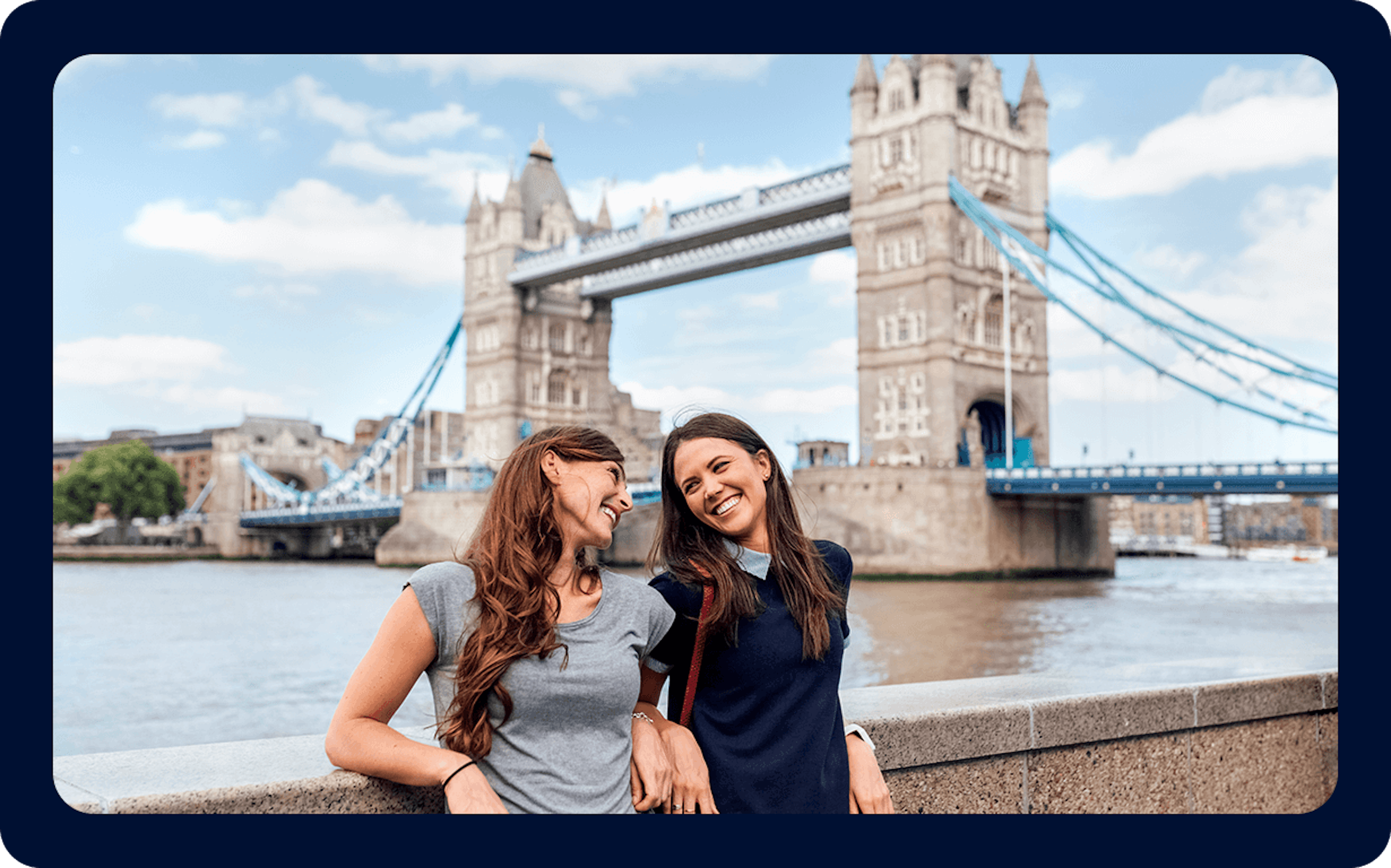 Dos mujeres sonrientes se encuentran junto a una barandilla de piedra con el Tower Bridge al fondo en un día soleado. Una mujer tiene el pelo largo y castaño y lleva una camiseta gris, mientras que la otra tiene el pelo largo y oscuro y lleva una camiseta azul marino. El puente, que aparece en muchos recursos de la "Guía para mudarse al Reino Unido", cruza un río tranquilo.