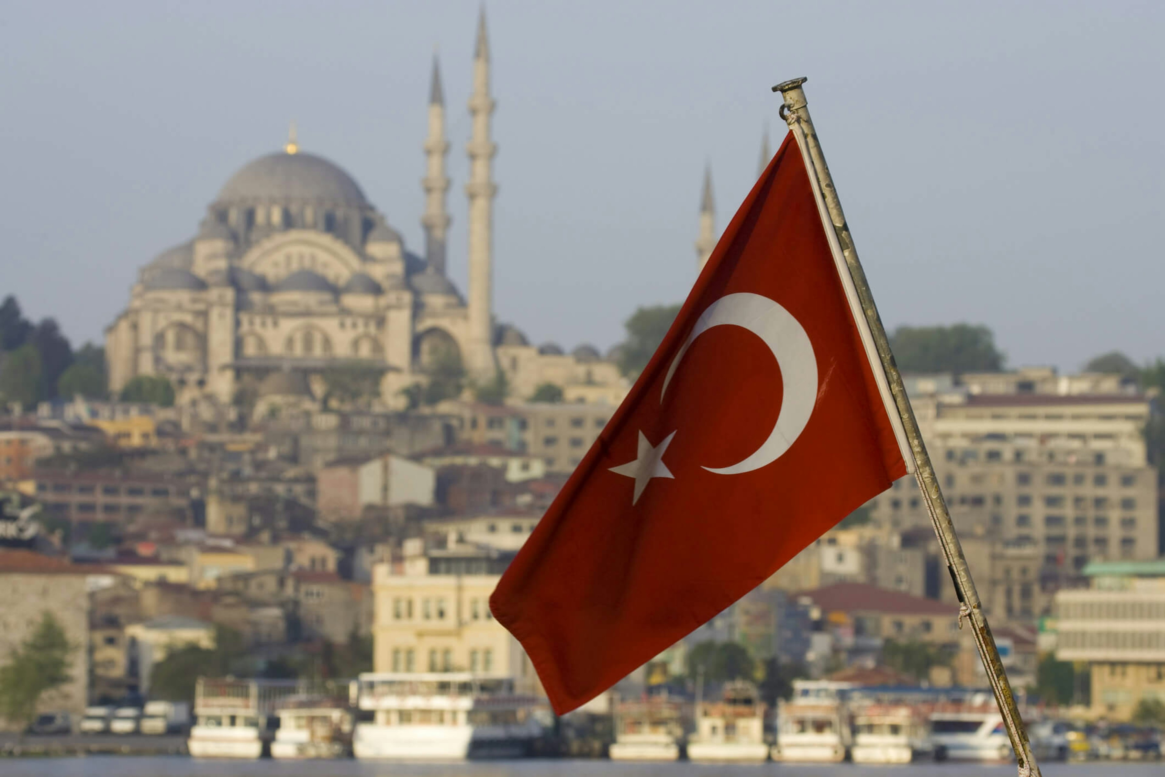 A Turkish flag flutters in the foreground, with the cityscape of Istanbul and the prominent Süleymaniye Mosque visible in the background under a clear sky. The scene includes boats on the water and historic buildings along the shoreline.