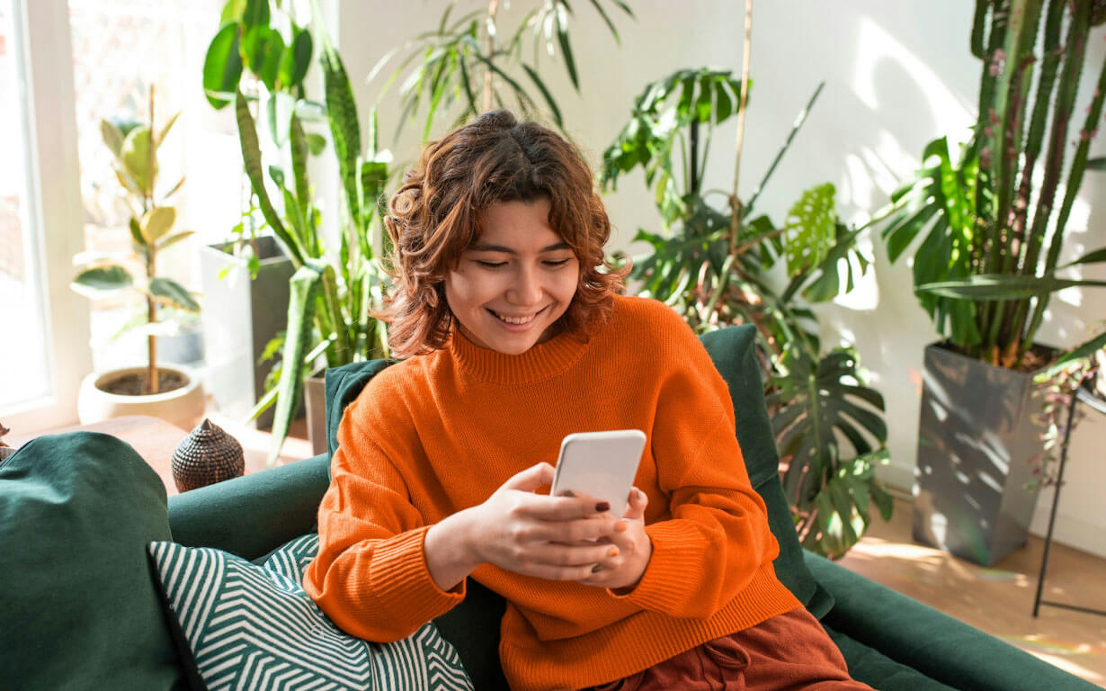 A person with curly hair, wearing an orange sweater, is sitting on a green couch while smiling at their smartphone. The background features various indoor plants with light streaming through the window.