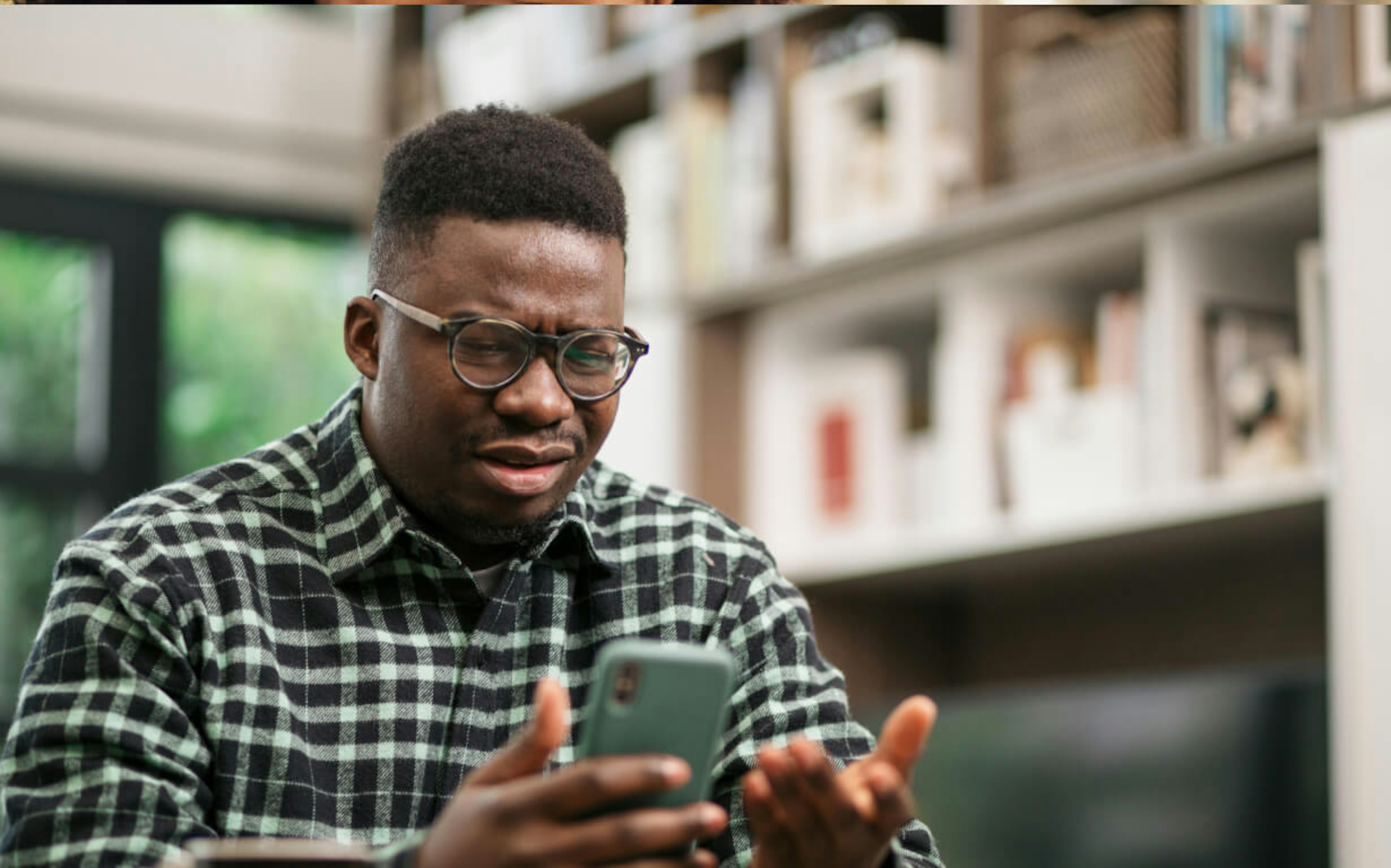 Un homme en chemise à carreaux est assis à l'intérieur, les yeux rivés sur son smartphone, l'air perplexe, peut-être intrigué par le dernier taux de change. Une main fait un geste devant lui comme pour essayer de résoudre l'énigme. Derrière lui, des étagères remplies de livres et d'objets de décoration ajoutent une touche chaleureuse à l'ensemble.