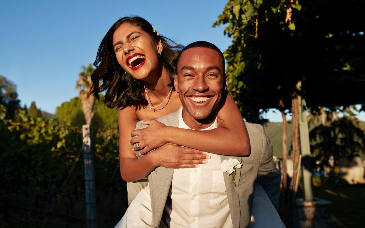 In a picture-perfect vineyard setting, a joyful couple captures the essence of Valentine's Day. The woman, in a white dress, playfully rides on her partner's back, both laughing under the clear blue sky. The man, dressed in a light gray suit, smiles warmly at the camera amidst lush greenery.