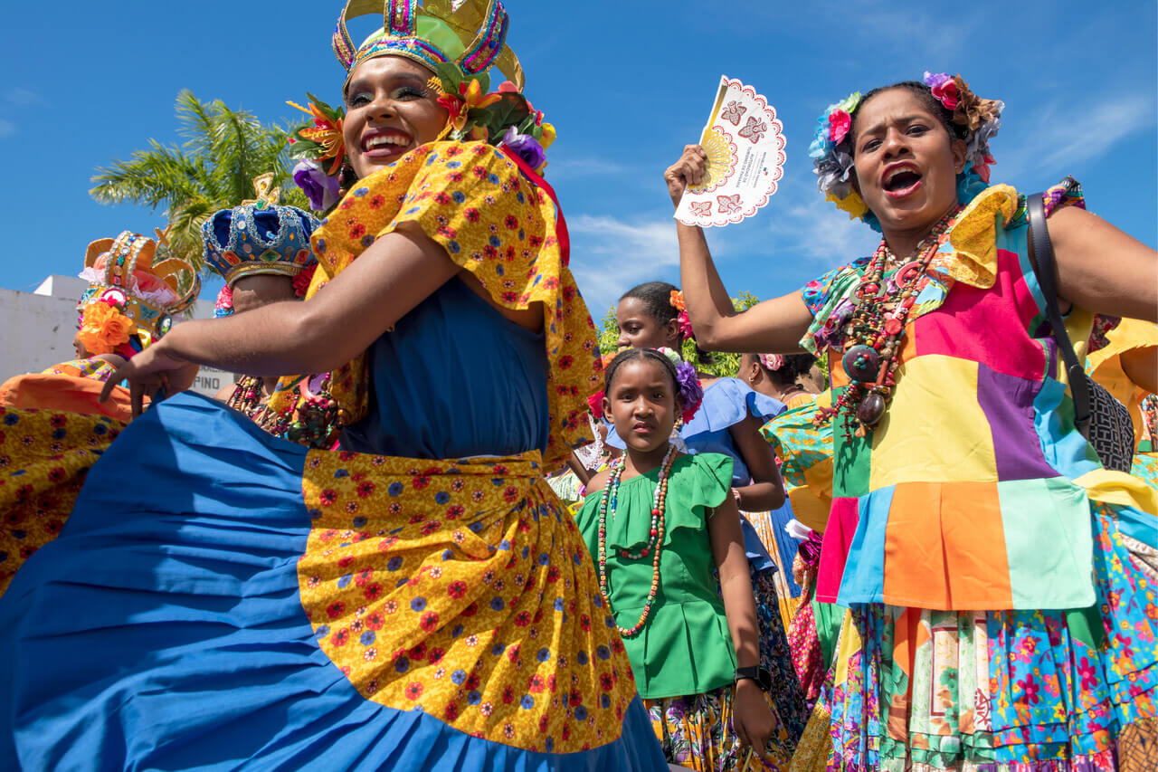 A group of women and girls in colorful, traditional Panamanian dresses dance and celebrate outdoors under a bright blue sky. The women smile joyfully, wearing vibrant floral headpieces and beaded necklaces.